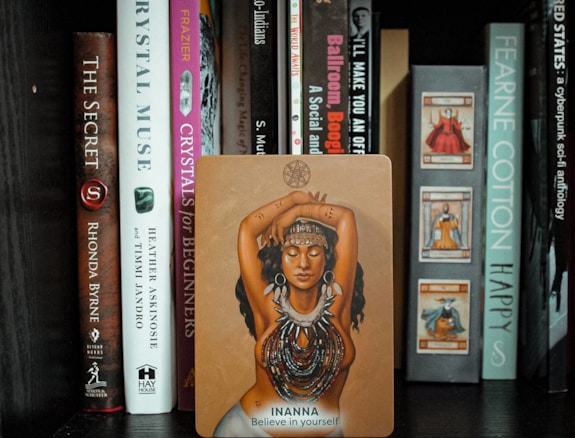 A shelf filled with various books, featuring a prominent card in the center depicting a woman with her arms crossed above her head, adorned with necklaces and symbols. The card reads 'Inanna - Believe in yourself.'