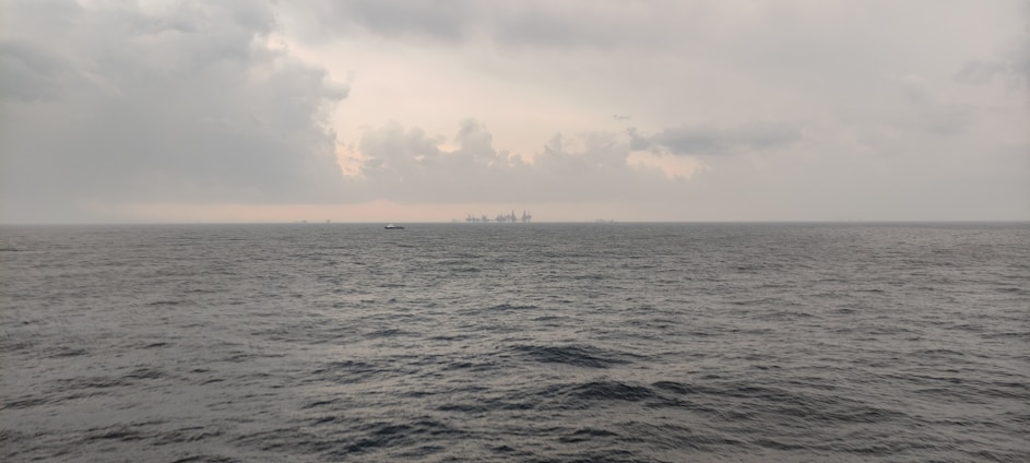 A vast expanse of ocean under a cloudy sky, with a distant view of an offshore oil rig and a ship on the horizon. The sea appears to be relatively calm despite the overcast weather.