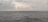 A vast expanse of ocean under a cloudy sky, with a distant view of an offshore oil rig and a ship on the horizon. The sea appears to be relatively calm despite the overcast weather.
