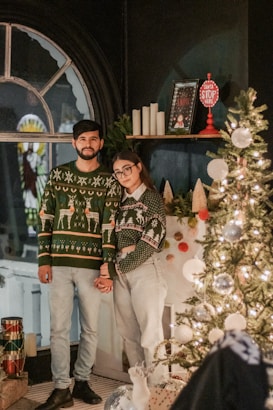 A man and woman stand side by side in a festive setting, both wearing Christmas-themed sweaters and light-colored jeans. Behind them, a decorated Christmas tree with lights and ornaments adds to the holiday spirit. Nearby, holiday decor including candles and a sign reading 'Santa Stop Here' is placed on a shelf. The room features a large arched window and the atmosphere is cozy and warm.