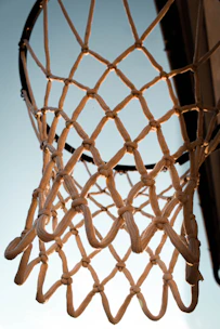 Close-up of a handwoven white basketball net hanging from a rusty outdoor hoop under a bright blue sky.