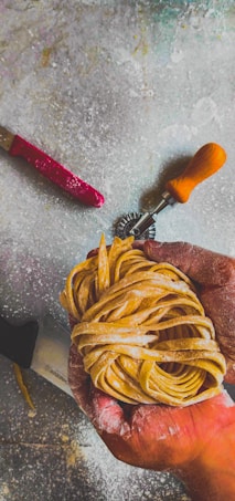 A hand holding a bundle of freshly made pasta, with a dusting of flour on the surface. Nearby tools include a red-handled knife and a pastry wheel with a wooden handle.