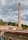 A tall brick chimney rises prominently against a cloudy sky. Surrounding the chimney are industrial buildings with brick and stone walls, topped with various pipes and antennas. In the background, there are trees and foliage providing a green backdrop.