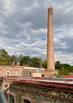 A tall brick chimney rises prominently against a cloudy sky. Surrounding the chimney are industrial buildings with brick and stone walls, topped with various pipes and antennas. In the background, there are trees and foliage providing a green backdrop.