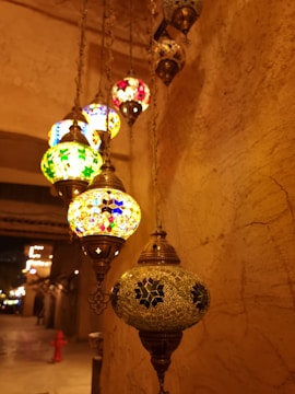A line of intricately designed hanging lanterns with colorful glass mosaics. The lanterns emit warm, vibrant light, casting artistic shadows on the stone walls. The background is dimly lit, resembling a traditional market setting.