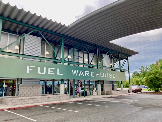 A commercial building with a sign reading 'FUEL WAREHOUSE'. It features a green and gray exterior with large windows and an overhanging roof. The building is located in a parking lot with visible trees and cars in the background.