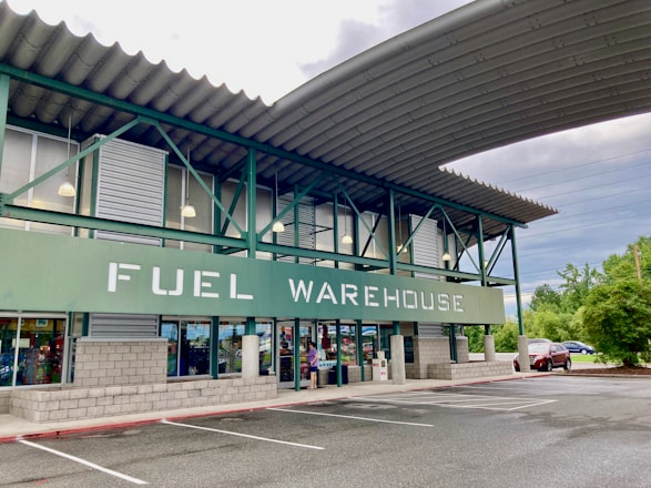 A commercial building with a sign reading 'FUEL WAREHOUSE'. It features a green and gray exterior with large windows and an overhanging roof. The building is located in a parking lot with visible trees and cars in the background.