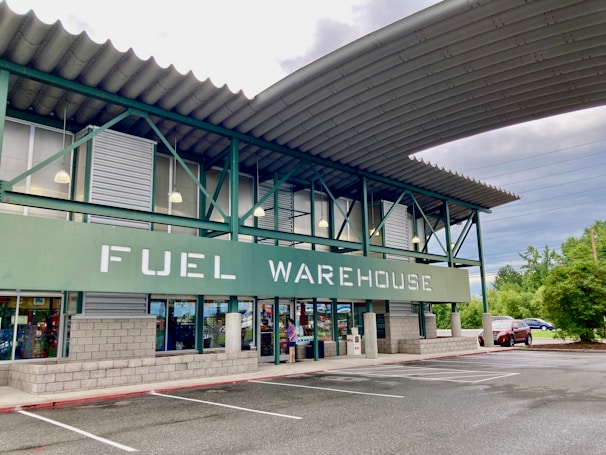 A commercial building with a sign reading 'FUEL WAREHOUSE'. It features a green and gray exterior with large windows and an overhanging roof. The building is located in a parking lot with visible trees and cars in the background.