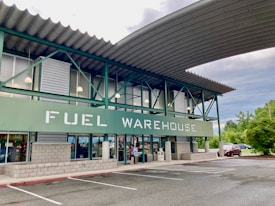 A commercial building with a sign reading 'FUEL WAREHOUSE'. It features a green and gray exterior with large windows and an overhanging roof. The building is located in a parking lot with visible trees and cars in the background.