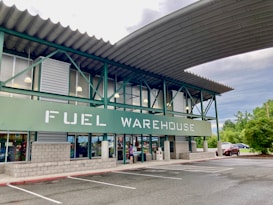 A commercial building with a sign reading 'FUEL WAREHOUSE'. It features a green and gray exterior with large windows and an overhanging roof. The building is located in a parking lot with visible trees and cars in the background.
