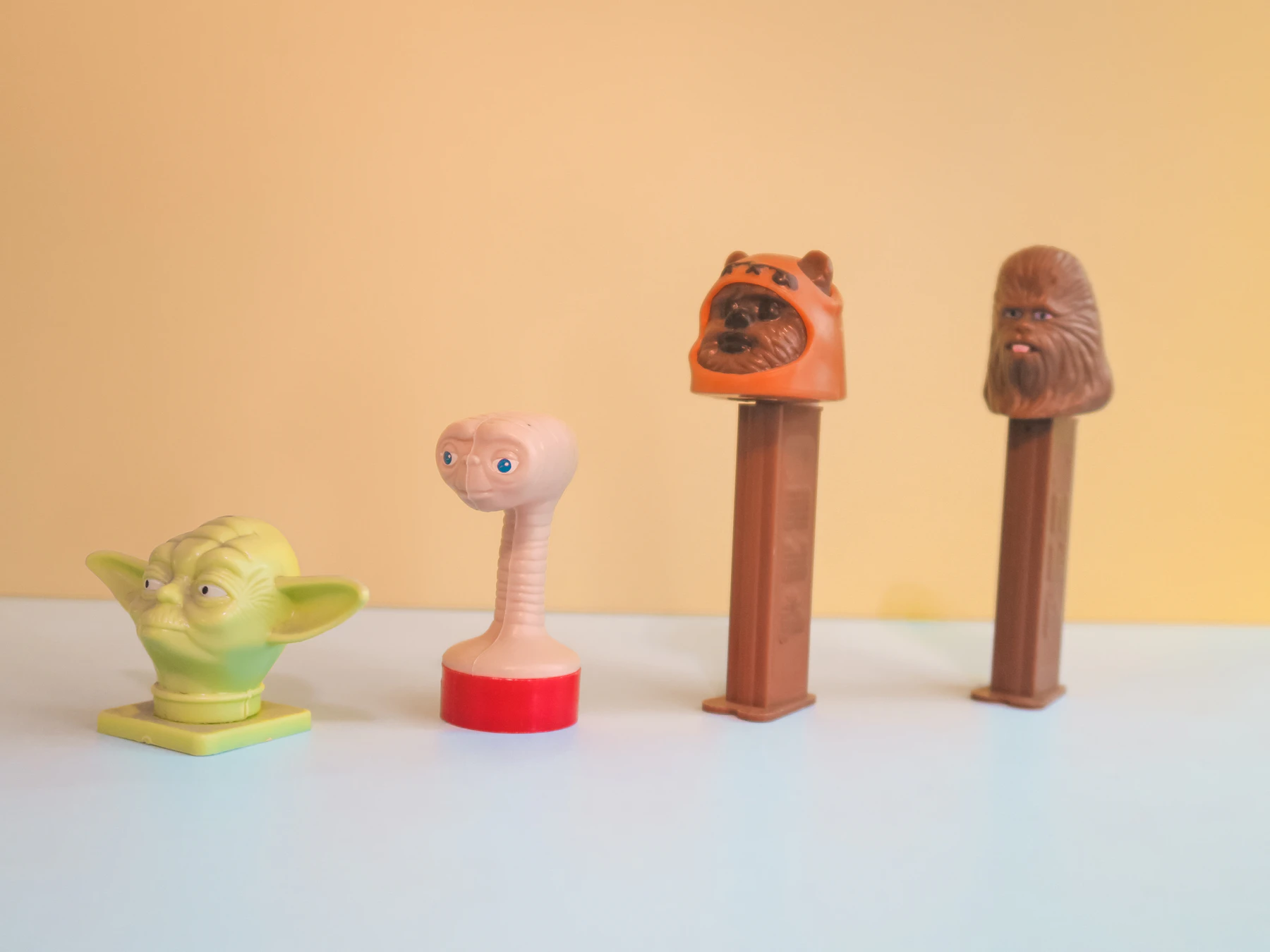 Two Pez dispensers with brown character heads standing next to two small toy figures against a yellow wall