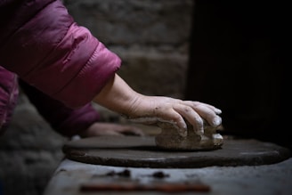 A hand pressing down on a block of clay, with the person wearing a maroon padded jacket. The scene is indoors with a dark, muted background, suggesting a workshop or studio environment.