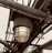 A close-up view of an industrial-style light fixture with a metal cage around the bulb. The fixture is mounted on a series of metal pipes and beams, showing signs of rust and wear. The background is overcast, adding a moody atmosphere to the scene.