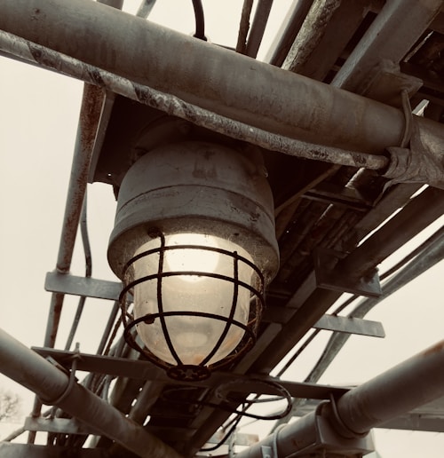 A close-up view of an industrial-style light fixture with a metal cage around the bulb. The fixture is mounted on a series of metal pipes and beams, showing signs of rust and wear. The background is overcast, adding a moody atmosphere to the scene.