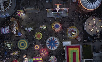 An overhead view of a busy amusement park featuring a variety of colorful carnival rides and booths. Circular and rectangular tents, along with spinning rides, are prominent amid a bustling crowd.