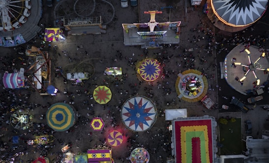 An overhead view of a busy amusement park featuring a variety of colorful carnival rides and booths. Circular and rectangular tents, along with spinning rides, are prominent amid a bustling crowd.