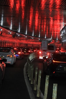 A tunnel filled with vehicles, predominantly cars and a truck, under a ceiling lined with reflective red lights. Lines of reflective barriers separate the lanes as traffic moves through the dimly lit passage.