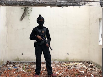 A person dressed in tactical gear, including a black helmet and face covering, stands in a room with broken, exposed walls and a floor covered in debris and bricks. The room has a rough, unfinished appearance with a partially open ceiling showing some metal beams and concrete.