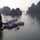 Boats are anchored in a scenic bay surrounded by large limestone karsts rising from the water. The serene water reflects the overcast sky, creating a tranquil scene. Several tour boats and smaller vessels are visible, suggesting a popular tourist destination.