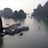 Boats are anchored in a scenic bay surrounded by large limestone karsts rising from the water. The serene water reflects the overcast sky, creating a tranquil scene. Several tour boats and smaller vessels are visible, suggesting a popular tourist destination.