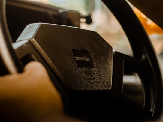 A steering wheel with a visible Nissan logo is held by a hand inside a car. The focus is on the center of the steering wheel, which is dark with a textured finish. There is a blurred view of the outside environment through the car window.