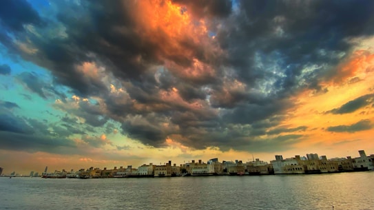 A dramatic sky with vibrant hues of orange and blue, dominated by thick, dark clouds. Below is a serene body of water reflecting the buildings that line its edge, showcasing a silhouette of an urban skyline.