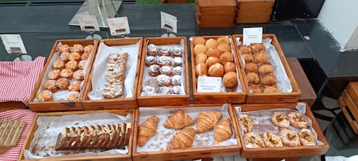 A display of various pastries on a buffet table, with different types of baked goods arranged in wooden trays. The pastries include flaky croissants, rolls dusted with powdered sugar, rectangular puff pastries, spherical bread rolls, and danish pastries. The table is covered with a red and white checkered cloth and labeled name cards are visible above the trays.