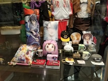 A display window featuring an assortment of anime-themed merchandise, including character pillows, mugs, hats, jackets, and figurines. The items showcase popular anime characters and are arranged neatly in a store setting.