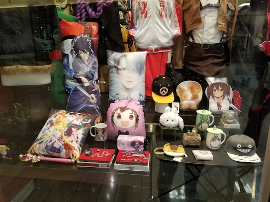 A display window featuring an assortment of anime-themed merchandise, including character pillows, mugs, hats, jackets, and figurines. The items showcase popular anime characters and are arranged neatly in a store setting.