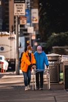 Two individuals, both wearing masks and casual clothing, are walking on a city sidewalk. One person is assisting the other who is using a walker. The scene includes urban elements such as signage, vehicles, and a trash bin.