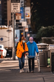 Two individuals, both wearing masks and casual clothing, are walking on a city sidewalk. One person is assisting the other who is using a walker. The scene includes urban elements such as signage, vehicles, and a trash bin.
