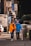 Two individuals, both wearing masks and casual clothing, are walking on a city sidewalk. One person is assisting the other who is using a walker. The scene includes urban elements such as signage, vehicles, and a trash bin.