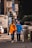 Two individuals, both wearing masks and casual clothing, are walking on a city sidewalk. One person is assisting the other who is using a walker. The scene includes urban elements such as signage, vehicles, and a trash bin.