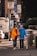 Two individuals, both wearing masks and casual clothing, are walking on a city sidewalk. One person is assisting the other who is using a walker. The scene includes urban elements such as signage, vehicles, and a trash bin.