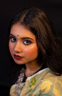 A portrait of a woman with long dark hair, wearing intricate makeup including bold eyeliner and deep red lipstick. She has a small black bindi on her forehead and is dressed in a traditional outfit with floral patterns and a necklace.