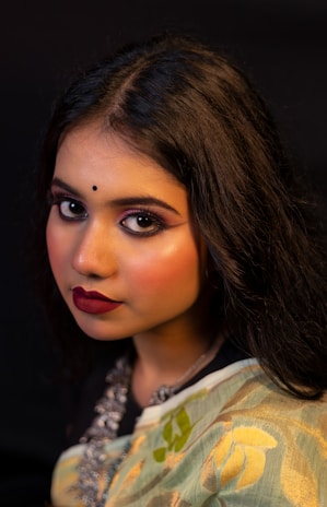 A portrait of a woman with long dark hair, wearing intricate makeup including bold eyeliner and deep red lipstick. She has a small black bindi on her forehead and is dressed in a traditional outfit with floral patterns and a necklace.