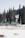 Several small cabins with green roofs are nestled amongst tall evergreen trees in a snowy landscape. The cabins are surrounded by a fence, and snow covers the ground, with patches of bare earth visible.