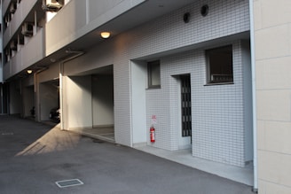 A modern building exterior with white tiled walls and windows. The area features a shadowy driveway or parking area under a row of small lights. There is a visible fire extinguisher next to a wall. The setting is quiet, with no people present.