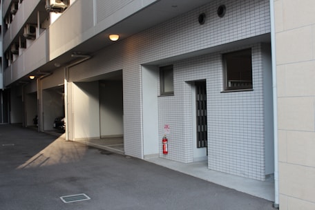 A modern building exterior with white tiled walls and windows. The area features a shadowy driveway or parking area under a row of small lights. There is a visible fire extinguisher next to a wall. The setting is quiet, with no people present.