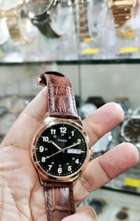 A hand is holding a wristwatch with a brown leather strap. The watch face is black with white numerals and has a date display. The background shows a display case with various other watches.