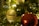 A close-up of a Christmas tree with a prominent round ornament featuring the word 'GRACE' engraved on it. The ornament hangs among green branches, with a blurred red bauble and glowing lights in the background.