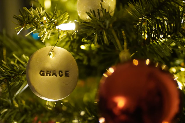 A close-up of a Christmas tree with a prominent round ornament featuring the word 'GRACE' engraved on it. The ornament hangs among green branches, with a blurred red bauble and glowing lights in the background.