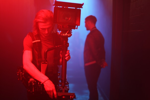 A person is holding a large, professional camera setup with stabilizing equipment in a dimly lit environment. The lighting in the scene features strong red and blue hues, casting dramatic shadows. In the background, another person stands with their back slightly turned.