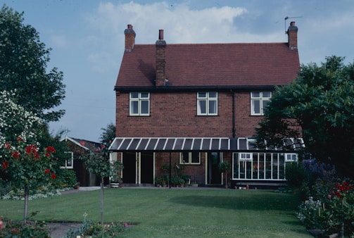 A two-story brick house with a red tiled roof sits amid a well-maintained garden. The house features multiple windows and a glass-enclosed veranda. Lush greenery surrounds the house, including various flowering plants and trees.