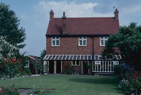 A two-story brick house with a red tiled roof sits amid a well-maintained garden. The house features multiple windows and a glass-enclosed veranda. Lush greenery surrounds the house, including various flowering plants and trees.