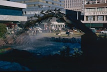 A public square features a water fountain with a sculpture resembling a tree or a series of branches arcing over the flowing water. Surrounding the fountain are green spaces with people relaxing on benches and grass. In the background, mid-century modern buildings with large glass windows and signs are visible, adding to the urban atmosphere.
