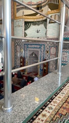 A richly decorated interior featuring intricate Islamic patterns and calligraphy on the walls. The room includes a mihrab, indicating a mosque setting, and a group of people seated on red carpeted flooring. Ornamental elements adorn the walls, and there's a mural on the upper section showcasing religious imagery with vibrant colors.
