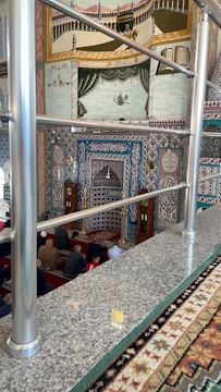 A richly decorated interior featuring intricate Islamic patterns and calligraphy on the walls. The room includes a mihrab, indicating a mosque setting, and a group of people seated on red carpeted flooring. Ornamental elements adorn the walls, and there's a mural on the upper section showcasing religious imagery with vibrant colors.