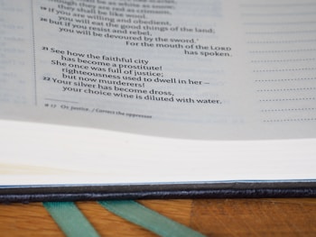 A close-up view of a book page featuring biblical verses. The text is focused and readable, detailing a passage with a somber tone. The page lies open on a wooden surface, and the book appears to have a dark leather binding.
