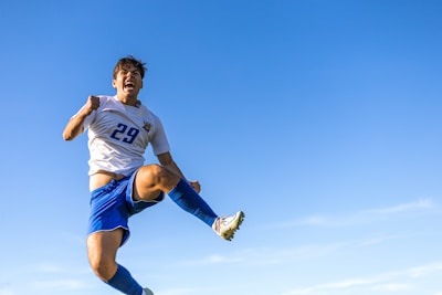 A soccer player is caught in mid-air against a clear blue sky, wearing a white jersey with the number 29 and blue shorts. The expression on the player's face suggests excitement or triumph, and the body posture indicates an athletic jump or kick.